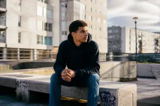 Jeune homme pensif en sweat à capuche assis sur un banc dans un quartier urbain, immeubles et tags en arrière-plan, lumière douce de fin de journée.