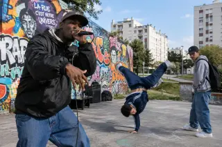 Un jeune homme rappe devant un mur de graffitis, accompagné par un danseur en plein mouvement et un spectateur, dans une rue de banlieue française sous une lumière de journée.