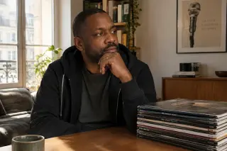 A Black man in his 40s with short hair and a trimmed beard, sitting at a table in a cozy urban living room, thoughtfully looking into the distance with a stack of eight vinyl records beside him and warm sunlight streaming through a window.